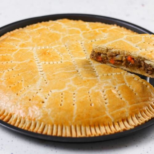 A large, golden-brown meat pie with a beautifully flaky crust, served on a round baking tray. One slice is lifted to reveal a hearty filling of minced beef, diced carrots, and potatoes inside. The crust features fork crimping around the edges and decorative vent holes across the surface.