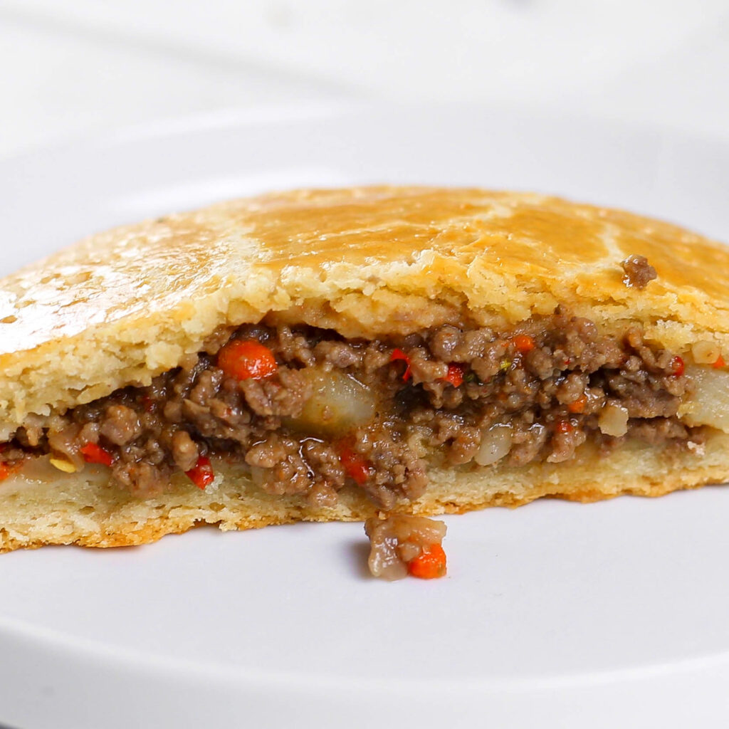 A close-up image of a meat pie slice, revealing its golden-brown, flaky crust and a filling of minced beef, diced potatoes, and red bell peppers. The juicy filling appears well-seasoned, with a slight sheen from the cooking juices. The pie is presented on a white plate, highlighting its appetizing texture and vibrant colors.