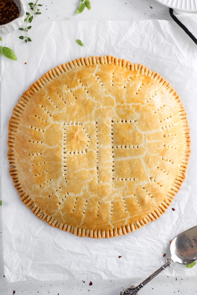 A beautifully baked golden-brown giant meat pie with “PIE” decoratively vented on the top crust. The pie is round with crimped edges and small fork holes across the surface, giving it a rustic look. It is displayed on white parchment paper, surrounded by scattered herbs and a silver pie server nearby.