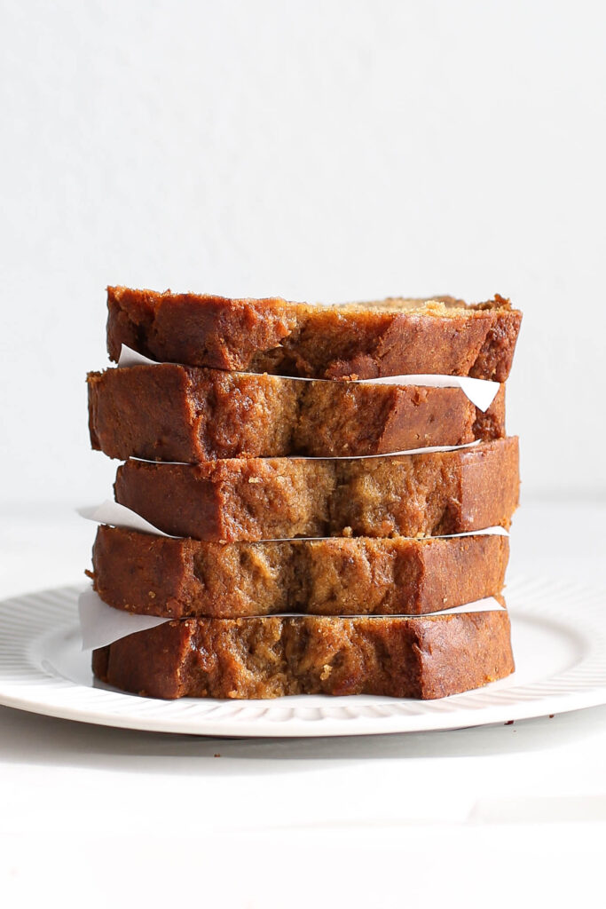 A stack of moist banana bread slices neatly arranged on a white plate. Each slice is separated by a small piece of parchment paper, showing off the golden-brown edges and soft, textured crumb. The bread looks perfectly baked, with hints of banana and spices visible throughout the loaf. The setting is simple and inviting, ideal for a cozy breakfast or snack.