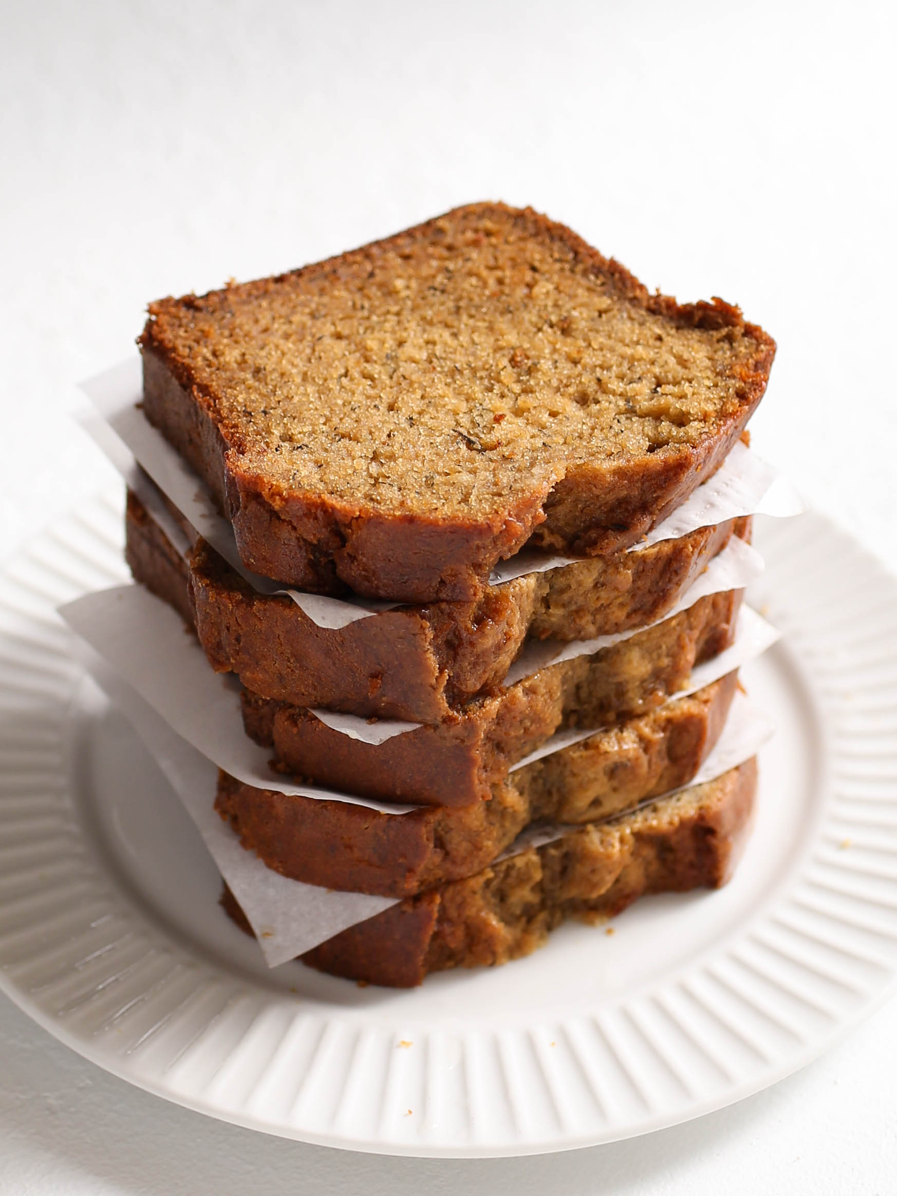 A stack of moist banana bread slices neatly arranged on a white plate. Each slice is separated by a small piece of parchment paper, showing off the golden-brown edges and soft, textured crumb. The bread looks perfectly baked, with hints of banana and spices visible throughout the loaf. The setting is simple and inviting, ideal for a cozy breakfast or snack.