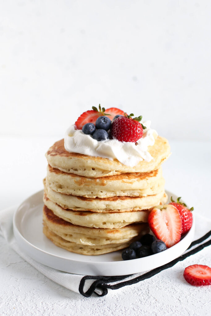  A tall stack of fluffy pancakes sits on a white plate, topped with a dollop of whipped cream, fresh strawberries, and blueberries. Additional berries are scattered around the base of the pancakes, along with a halved strawberry for garnish. The pancakes are golden and thick, highlighting their light, airy texture. The background is minimal and bright, drawing focus to the colorful, delicious-looking pancakes. A black and white napkin is placed beneath the plate.