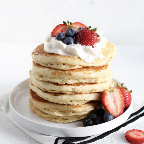 A tall stack of fluffy pancakes sits on a white plate, topped with a dollop of whipped cream, fresh strawberries, and blueberries. Additional berries are scattered around the base of the pancakes, along with a halved strawberry for garnish. The pancakes are golden and thick, highlighting their light, airy texture. The background is minimal and bright, drawing focus to the colorful, delicious-looking pancakes. A black and white napkin is placed beneath the plate.