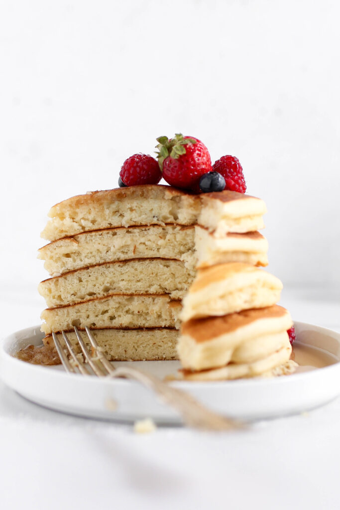 A stack of fluffy pancakes is topped with fresh strawberries, raspberries, and blueberries. A fork is placed next to a bite-sized section of the pancakes, revealing their soft, airy texture. The pancakes sit on a white plate, and maple syrup can be seen pooling beneath the stack. The background is clean and minimal, highlighting the golden-brown pancakes.