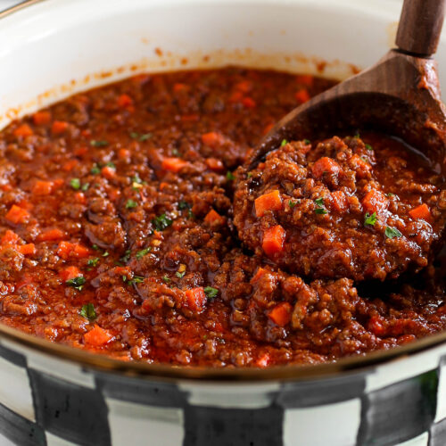 A close-up image of a pot filled with a rich, chunky minced meat sauce. The sauce is a deep red color with visible bits of minced meat, diced carrots, and a hint of green herbs for garnish. A wooden spoon is lifting a portion of the sauce, showing its thick, hearty texture. The pot has a black-and-white checkered pattern along its rim, giving a rustic feel to the presentation.