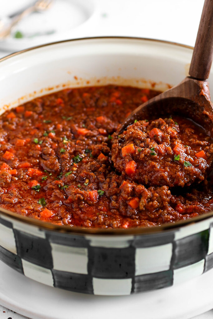 A close-up image of a pot filled with a rich, chunky minced meat sauce. The sauce is a deep red color with visible bits of minced meat, diced carrots, and a hint of green herbs for garnish. A wooden spoon is lifting a portion of the sauce, showing its thick, hearty texture. The pot has a black-and-white checkered pattern along its rim, giving a rustic feel to the presentation.