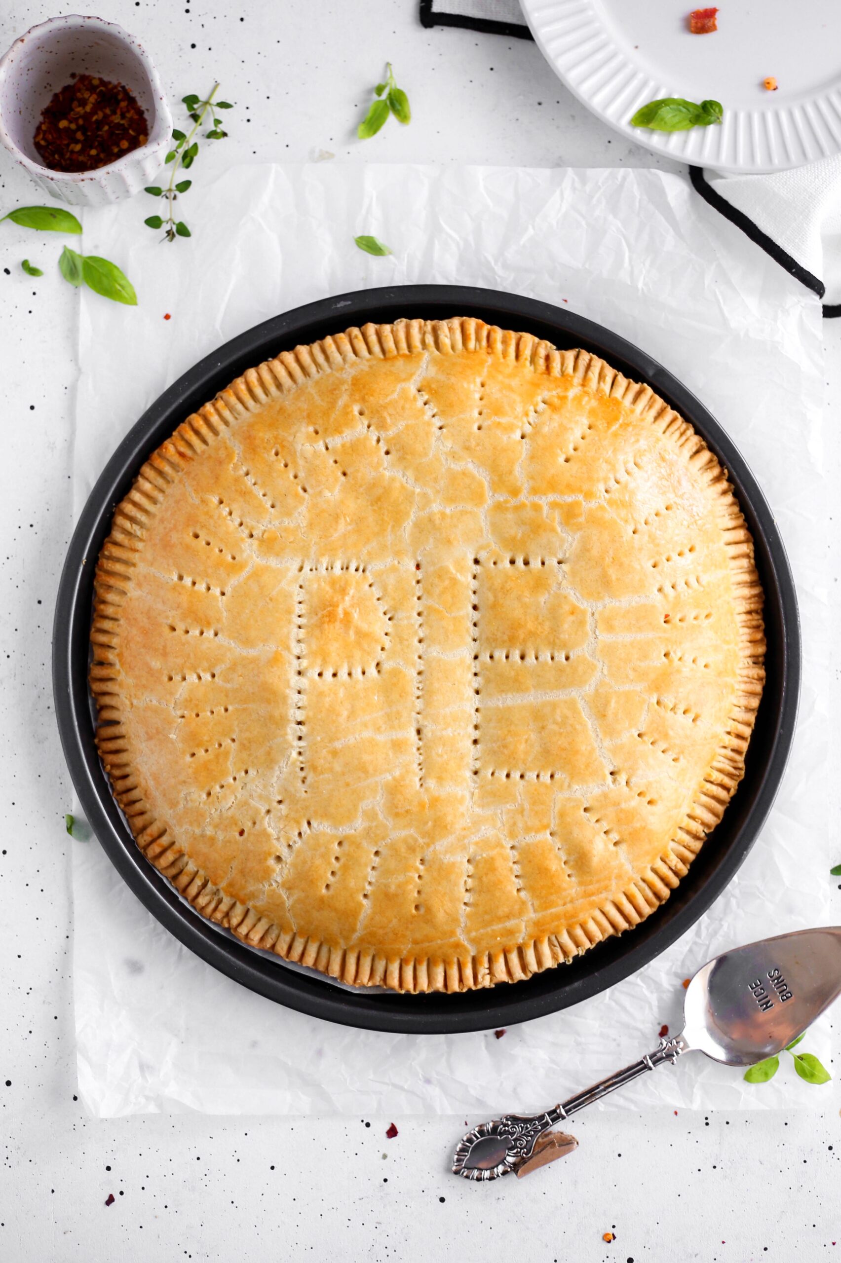 A beautifully baked golden-brown giant meat pie with “PIE” decoratively vented on the top crust. The pie is round with crimped edges and small fork holes across the surface, giving it a rustic look. It is displayed on white parchment paper, surrounded by scattered herbs and a silver pie server nearby.