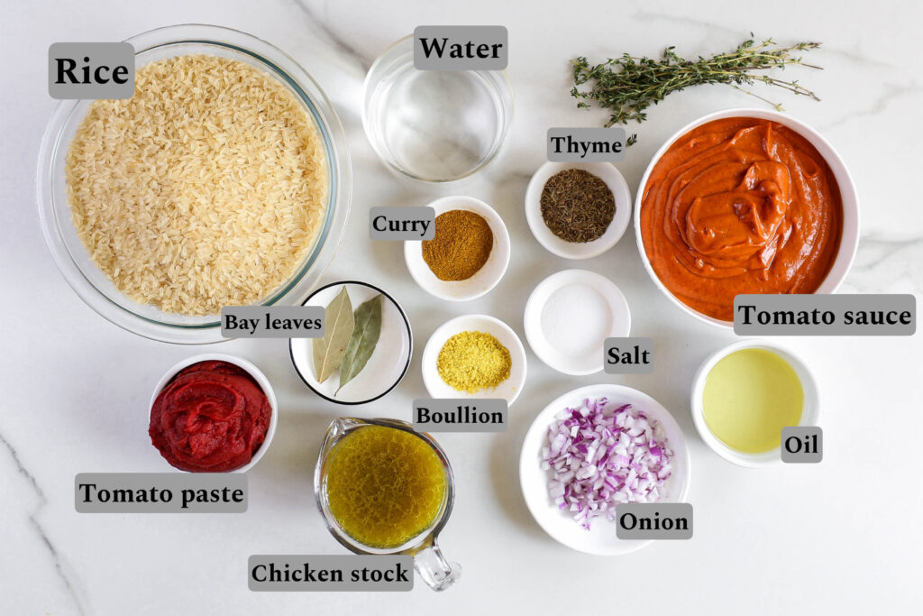 Alt text: A collection of ingredients for making Jollof rice arranged on a white countertop. The ingredients include a bowl of uncooked rice, a glass of water, a bunch of thyme sprigs, a bowl of tomato sauce, a small bowl of curry powder, a small bowl of dried thyme, a small bowl of salt, a bowl of oil, a small bowl of chopped red onions, a jug of chicken stock, a small bowl labeled “bouillon,” a bowl of tomato paste, and a small plate with bay leaves.